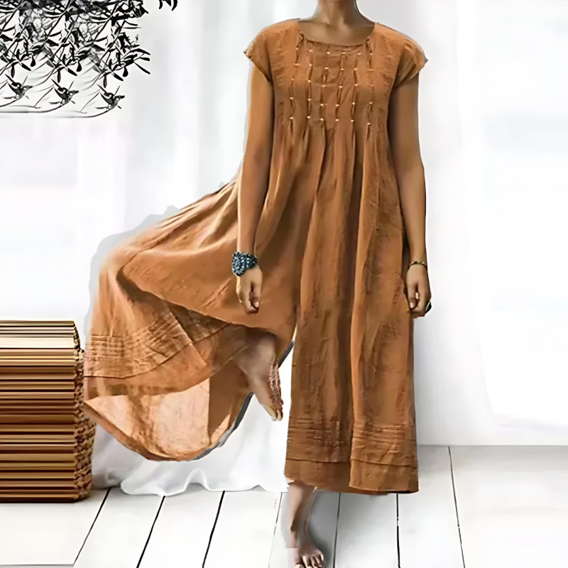 Femme portant une combinaison une-pièce couleur camel fluide, à manches courtes et coupe ample, avec de fins détails décoratifs sur le buste, réalisée dans un tissu léger et aérien tombant jusqu’aux chevilles, présentée dans un intérieur lumineux et épuré.