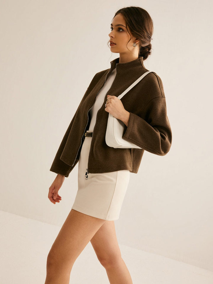 Femme marchant élégamment en portant un manteau court marron à col montant et fermeture zippée, assorti à une robe courte beige et un sac à main blanc, pour un look moderne et raffiné.