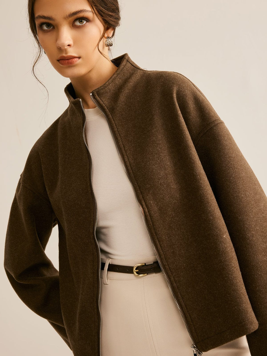 Femme portant un manteau court marron à col montant et fermeture zippée, assorti à une robe beige et une ceinture, pour un look moderne et raffiné.