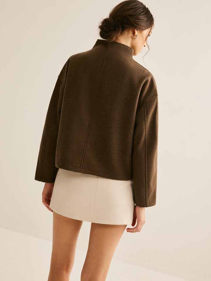 Vue arrière d'une femme portant un manteau court marron à col montant, avec une coupe moderne et une robe beige, créant un look élégant et raffiné.