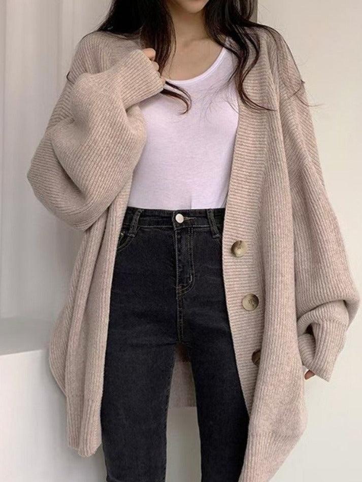 Femme portant un cardigan long en maille beige clair avec de grands boutons sur le devant. Le vêtement oversize est associé à un t-shirt blanc et un jean noir taille haute, offrant un look doux, moderne et décontracté idéal pour les journées d’automne ou d’hiver.