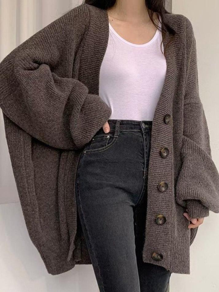 Une femme porte un long cardigan marron en maille, à gros boutons, à manches larges et à la coupe ample. Elle l'associe à un simple t-shirt blanc et un jean noir taille haute pour un look moderne, minimaliste et confortable, parfait pour la mi-saison.