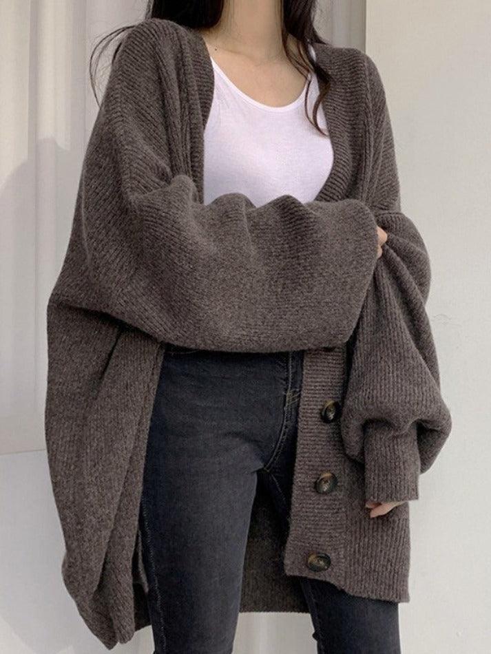 Une femme porte un long cardigan marron en maille, orné de gros boutons sur le devant. Ce vêtement ample est associé à un t-shirt blanc et un jean noir ajusté, créant ainsi un style moderne, confortable et minimaliste, idéal pour l'automne ou l'hiver.