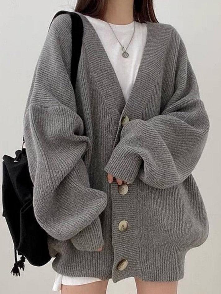 Femme portant un cardigan gris oversize en maille épaisse, avec de grands boutons sur le devant et des manches bouffantes. Le look est complété par un t-shirt blanc, un collier discret et un sac noir, pour un style décontracté et moderne idéal pour la mi-saison.
