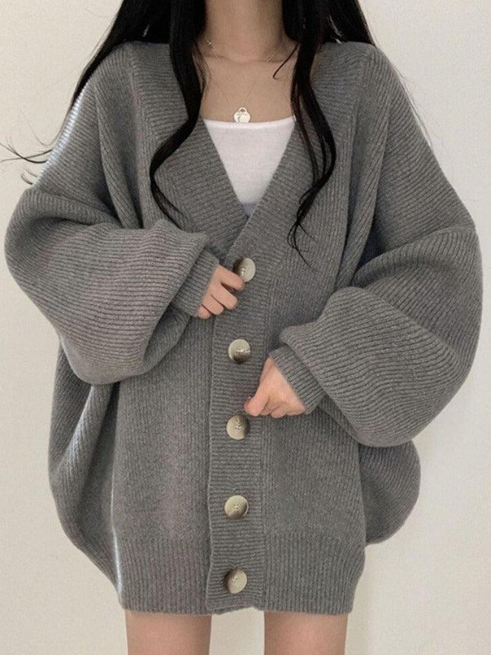 Femme portant un long cardigan gris en maille épaisse avec de grands boutons sur le devant. Le vêtement présente une coupe oversize et des manches amples, associé à un débardeur blanc, pour un style moderne, minimaliste et confortable adapté aux saisons fraîches.