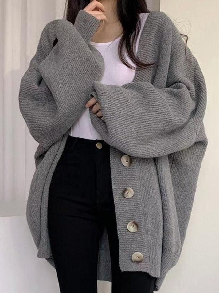 Femme portant un long cardigan gris en maille épaisse avec de grands boutons sur le devant. Le vêtement, au style ample et décontracté, est associé à un t-shirt blanc et un pantalon noir taille haute, créant un look moderne et confortable parfait pour les journées fraîches.