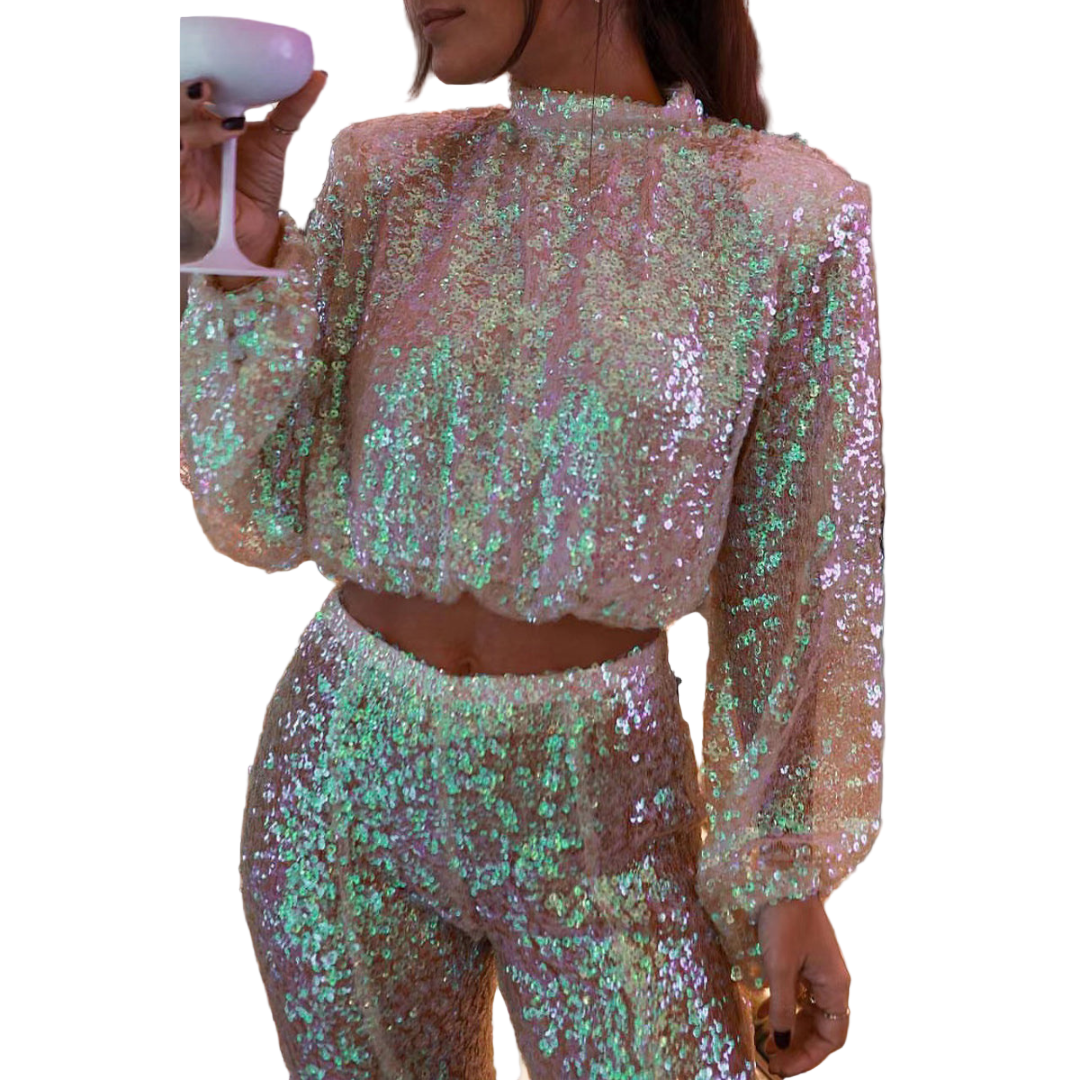 Femme portant un ensemble de soirée scintillant composé d’un haut à manches longues et d’un pantalon assorti, tous deux entièrement recouverts de sequins irisés. Le haut présente une coupe courte et légèrement bouffante avec un col montant, tandis que le pantalon taille haute affine la silhouette. La lumière se reflète sur les paillettes pour un effet élégant et glamour, idéal pour les fêtes et les événements de soirée.