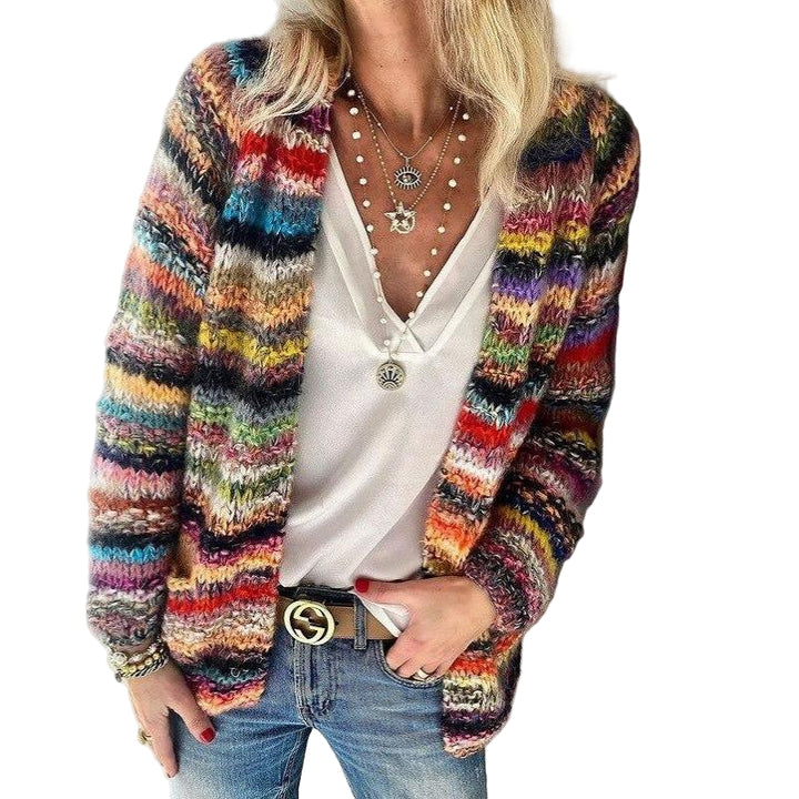 Femme portant un cardigan en maille multicolore à motifs rayés, associé à un haut blanc décolleté et un jean bleu. Elle porte plusieurs colliers superposés et un bracelet, avec une main dans la poche et l’autre posée le long du corps.