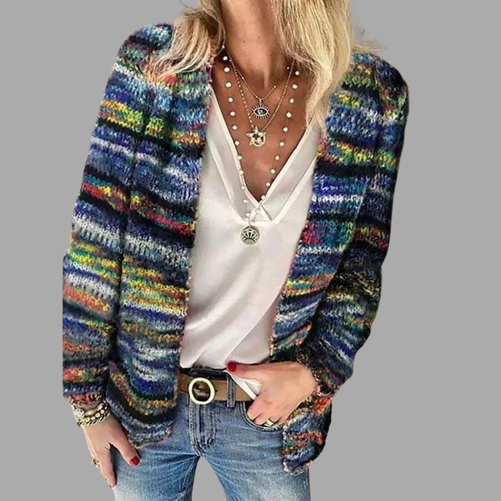 Femme portant un cardigan en maille multicolore dominé par des tons bleus, assorti à un haut blanc décolleté et un jean bleu. Elle porte plusieurs colliers superposés ainsi qu’un bracelet, avec une main dans la poche et l’autre reposant le long du corps.