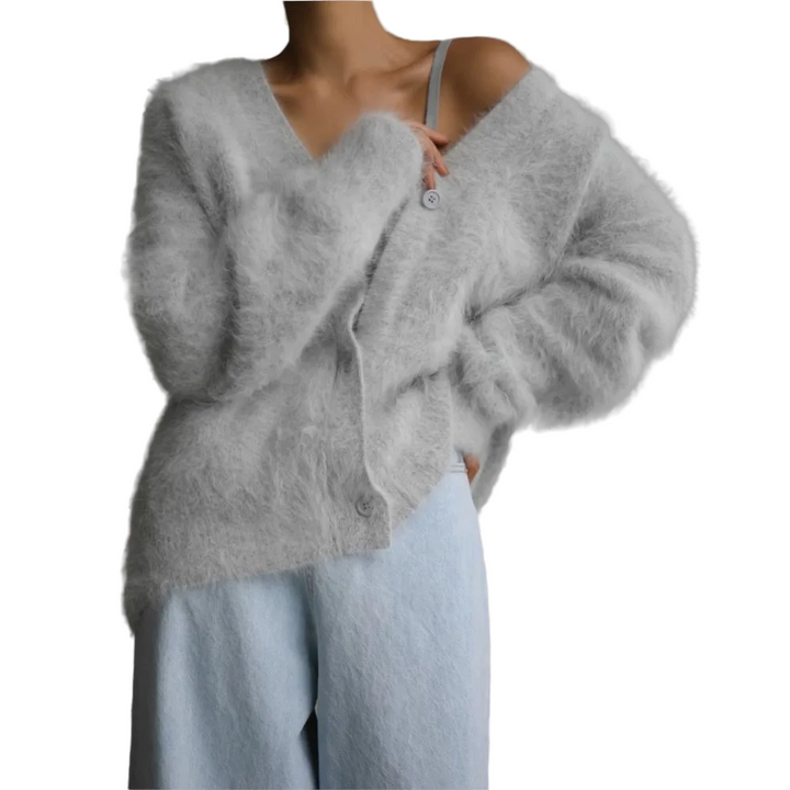 Femme portant un cardigan doux et duveteux de couleur gris clair avec un col en V et des boutons sur le devant, légèrement tombé sur une épaule, associé à un pantalon ample en laine pâle. L’ensemble dégage une allure moderne, confortable et élégante dans un cadre minimaliste.