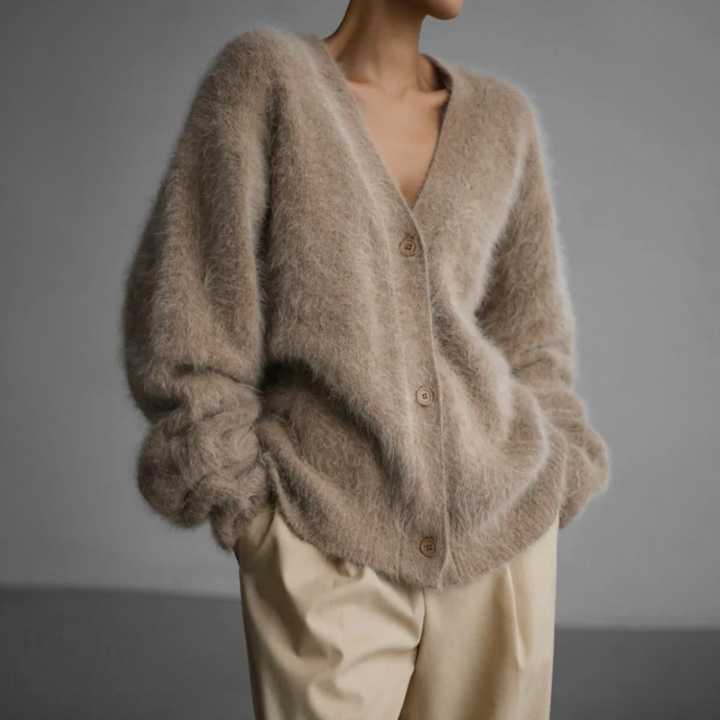 Femme portant un cardigan doux et moelleux de couleur beige clair, à col en V et à boutons sur le devant, associé à un pantalon ample de teinte crème. L’ensemble dégage une allure moderne, décontractée et élégante dans un cadre minimaliste.