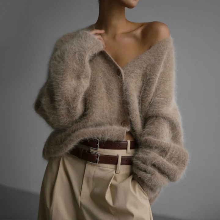 Femme portant un cardigan doux et duveteux de couleur beige, légèrement tombé sur une épaule, avec des boutons sur le devant. Le cardigan est rentré partiellement dans un pantalon beige à pinces, maintenu par une ceinture double en cuir marron. L’ensemble crée un style moderne, sensuel et raffiné dans un cadre minimaliste.