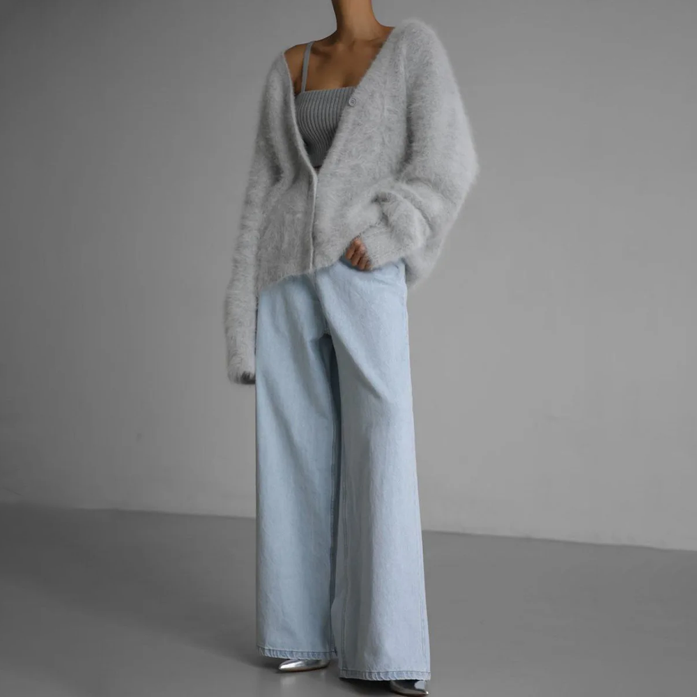 Femme portant un cardigan doux et duveteux de couleur gris clair avec un col en V, légèrement tombé sur une épaule, associé à un débardeur côtelé gris et un pantalon large en denim clair. L’ensemble dégage une allure moderne, confortable et sophistiquée dans un décor minimaliste.