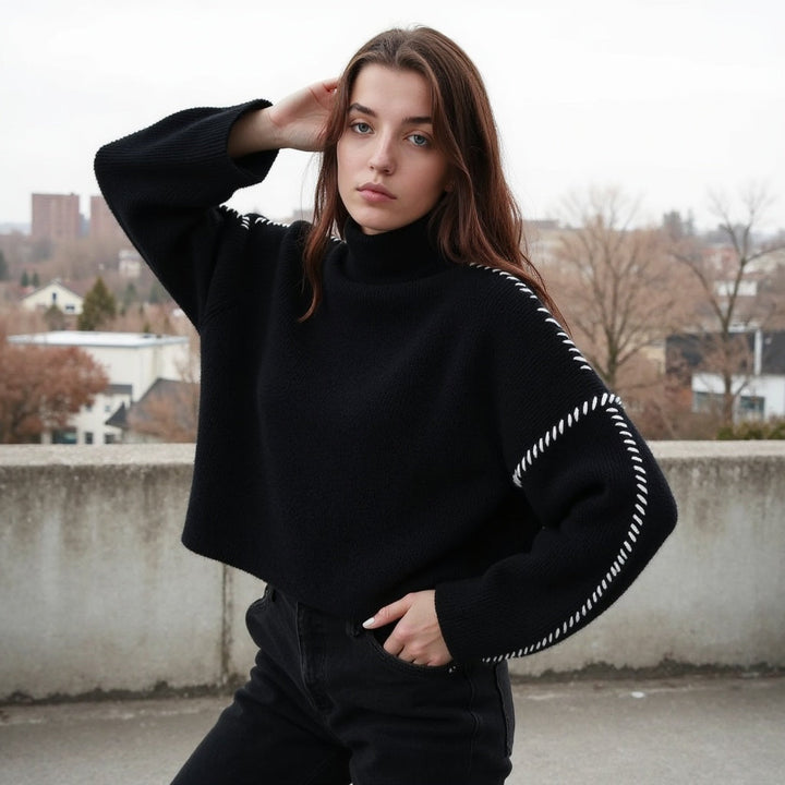 Jeune femme posant sur une terrasse, vêtue d’un pull noir à col montant avec des détails de couture blanche le long des manches. Elle a une main dans la poche et l’autre touchant ses cheveux. L’arrière-plan montre des arbres nus et des bâtiments résidentiels par temps gris.