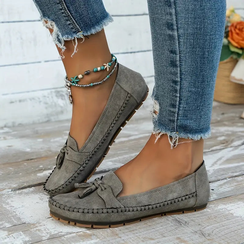 Vue latérale des mocassins plats gris pour femmes, avec un nœud décoratif et une finition en suède. Ces mocassins sont parfaits pour un look décontracté et moderne, complétés par des bracelets bohèmes et un jean déchiré. Confort et style assurés pour toutes les occasions.