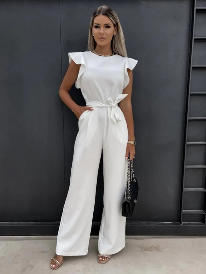 Combinaison blanche pour femme avec des manches volantées et une ceinture assortie à la taille. Elle présente une coupe large et élégante, complétée par une pochette noire et des sandales à talons, parfaite pour une occasion chic et moderne.