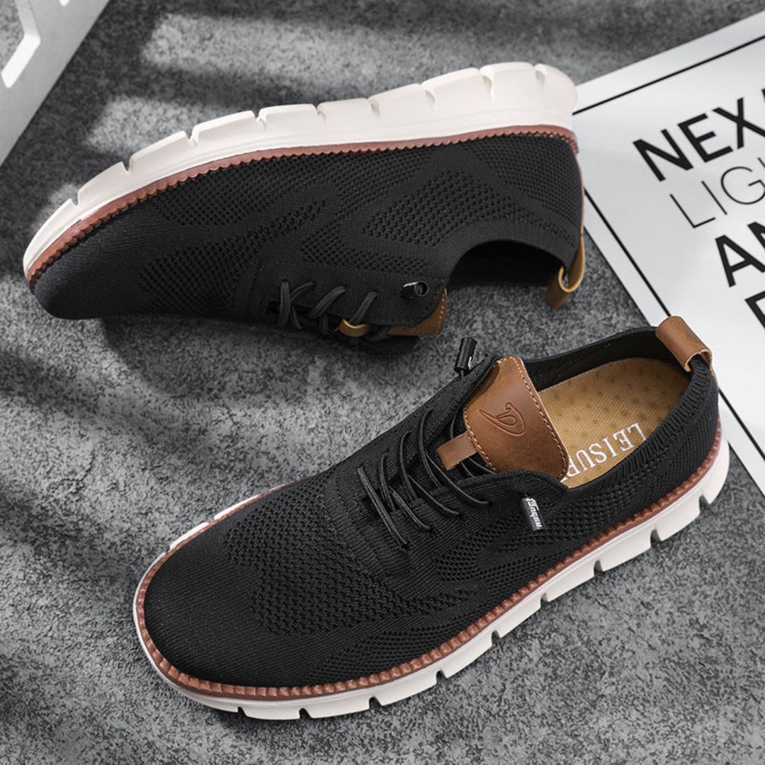 Paire de chaussures urbaines noires en maille respirante avec lacets, détails en simili cuir camel et semelle épaisse crantée blanche, posée sur un sol gris.