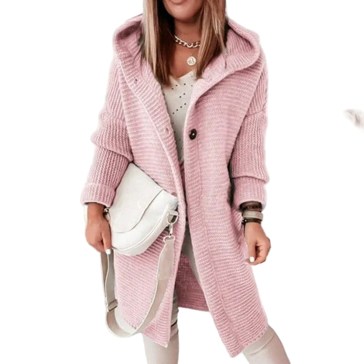 Femme portant un long cardigan en maille rose avec capuche, assorti à un pantalon clair et un sac bandoulière beige. Elle se tient debout dans un intérieur moderne décoré de portraits artistiques en arrière-plan.