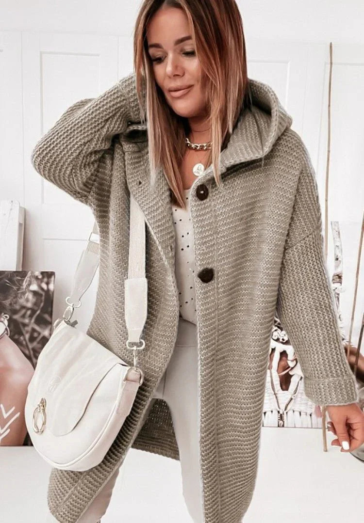 Une femme porte un long cardigan kaki à capuche, ouvert sur un haut clair, associé à un pantalon neutre. Elle tient un sac bandoulière crème et pose dans un intérieur lumineux orné de portraits artistiques.