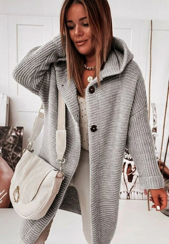Femme vêtue d’un long cardigan en maille gris clair avec capuche et gros boutons, porté sur un haut blanc à motifs discrets et un pantalon neutre. Elle tient un sac bandoulière crème et se tient dans un intérieur lumineux décoré de portraits artistiques.