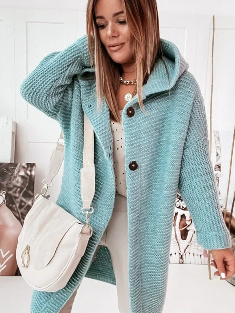 Une femme porte un long cardigan vert clair en maille, à capuche et gros boutons, associé à un haut blanc et un pantalon neutre. Elle tient un sac bandoulière crème et pose dans un intérieur lumineux orné de portraits artistiques.
