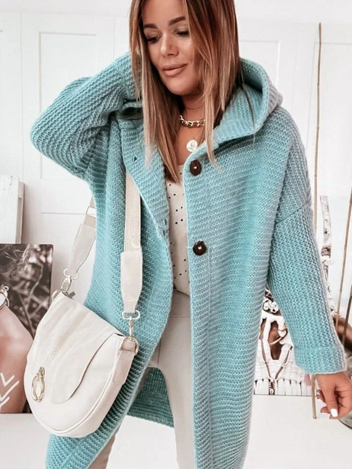 Une femme porte un long cardigan vert clair en maille, à capuche et gros boutons, associé à un haut blanc et un pantalon neutre. Elle tient un sac bandoulière crème et pose dans un intérieur lumineux orné de portraits artistiques.