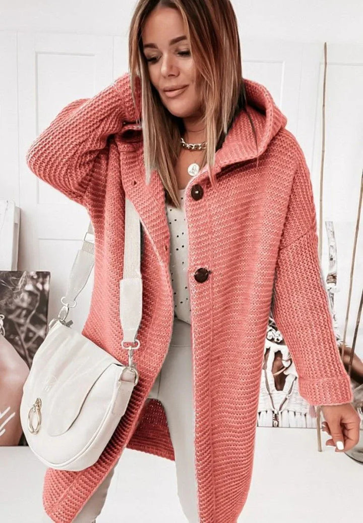 Une femme porte un long cardigan en maille rouge à capuche et gros boutons, associé à un haut blanc à petits motifs et un pantalon clair. Elle tient un sac bandoulière crème et pose dans un intérieur lumineux orné de portraits artistiques.