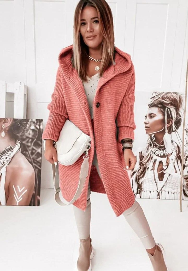 Une femme porte un long cardigan en maille rouge à capuche et gros boutons, associé à un haut blanc et un pantalon clair. Elle tient un sac bandoulière crème et pose dans un intérieur lumineux orné de portraits artistiques au sol et aux murs.