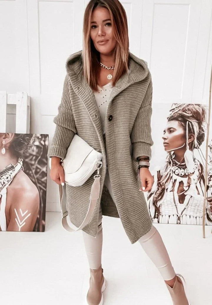 Une femme vêtue d'un long cardigan kaki en maille à capuche et gros boutons, porté sur un haut blanc et un pantalon clair, tient un sac à bandoulière crème et pose dans un intérieur lumineux orné de portraits artistiques disposés au sol et contre les murs.