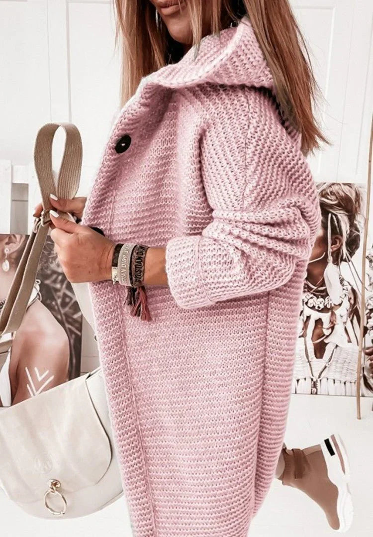 Femme de profil portant un long cardigan en maille rose pâle à texture épaisse, avec manches retroussées et gros boutons. Elle tient un sac bandoulière crème et porte plusieurs bracelets, posant dans un intérieur lumineux décoré de portraits artistiques.