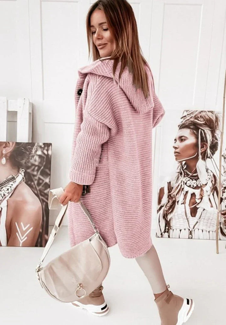 Femme vue de dos portant un long cardigan en maille rose avec capuche, assorti à un pantalon clair et des baskets beige et blanc. Elle tient un sac bandoulière crème et se trouve dans un intérieur lumineux décoré de portraits artistiques posés au sol et contre le mur.