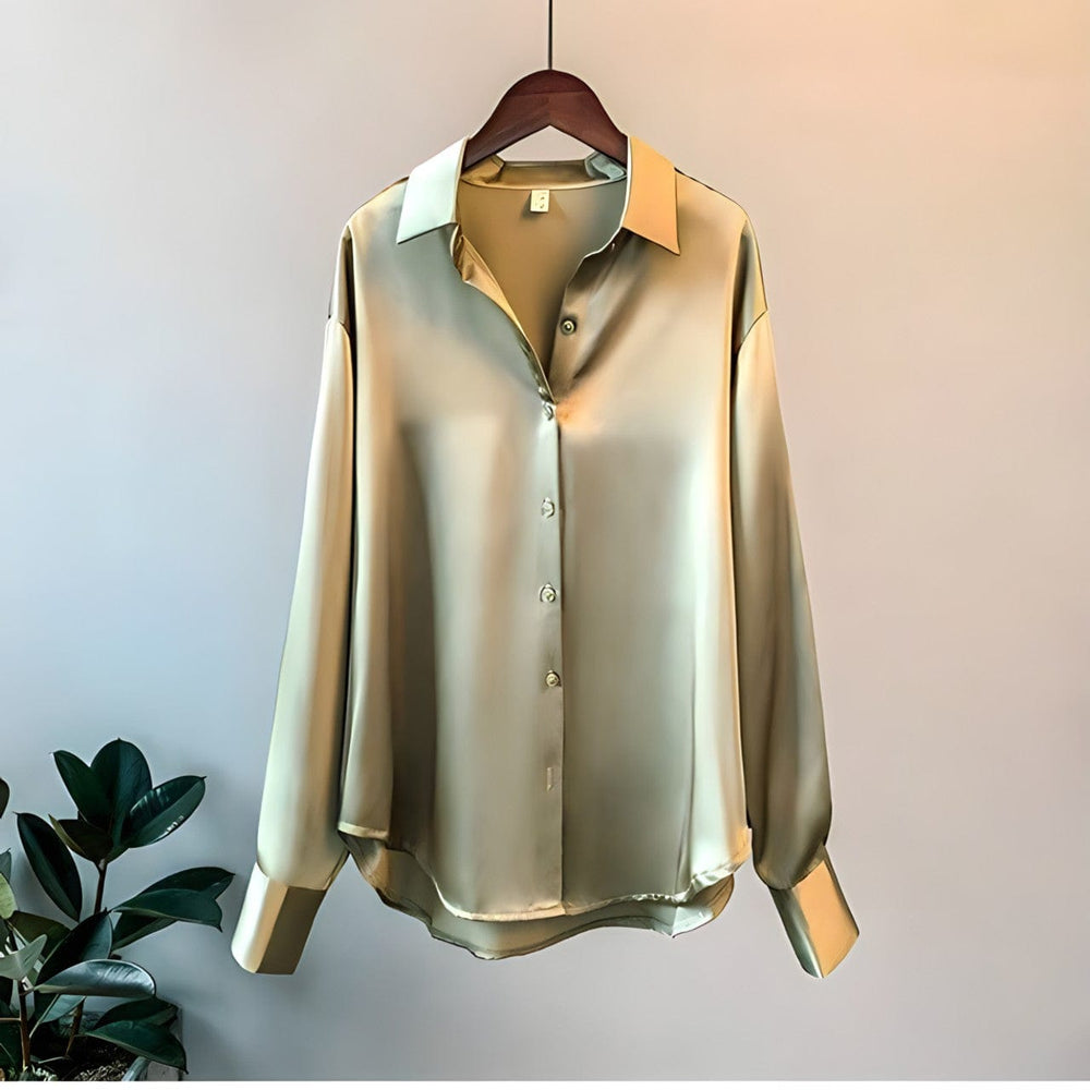 Chemise élégante à manches longues en satin de couleur dorée, suspendue sur un cintre en bois. Le tissu soyeux reflète la lumière pour un effet luxueux et fluide. Elle présente une coupe ample, un col classique et des boutons assortis, idéale pour un style raffiné et confortable au quotidien.