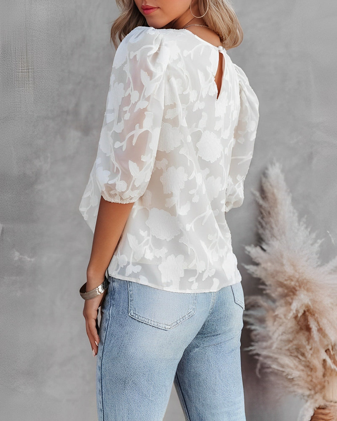 Vue arrière du chemisier pour femme, avec un imprimé floral ton sur ton sur fond blanc. Ce haut à manches bouffantes présente une ouverture délicate au dos, ajoutant une touche élégante à la coupe fluide. Le modèle le porte avec un jean décontracté, créant un look moderne et chic, parfait pour une tenue quotidienne stylée.