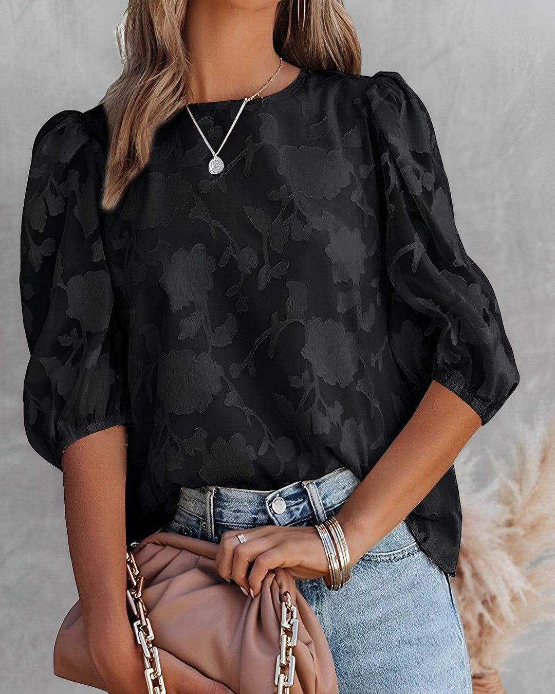 Chemisier pour femme, élégant et tendance, avec un imprimé floral discret sur fond noir. Ce haut à manches bouffantes offre une coupe moderne et chic, parfait pour des looks décontractés ou habillés. Le modèle porte le chemisier avec un jean et un sac élégant, créant une tenue à la fois stylée et confortable.