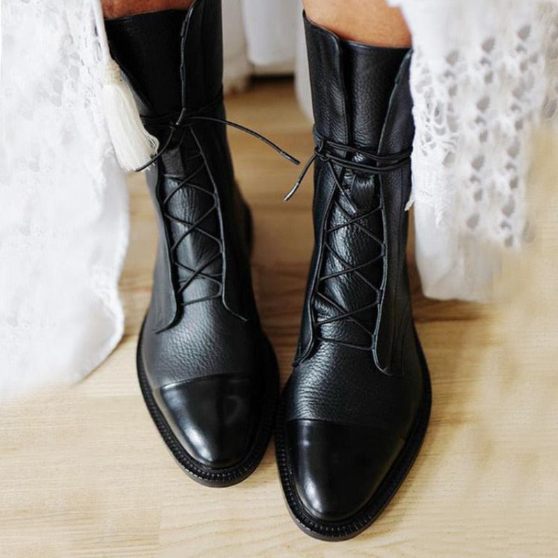 Bottines cheville pour femme en cuir noir à texture grainée, avec laçage fin sur le devant, bout arrondi et semelle plate robuste, portées avec une tenue claire en intérieur sur un sol en bois.