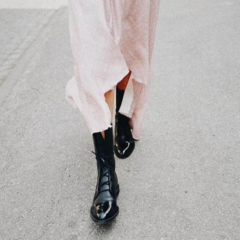 Bottines cheville pour femme en cuir noir à finition lisse et grainée, avec laçage sur le devant, bout arrondi et semelle plate robuste, portées en extérieur avec une robe fluide claire.