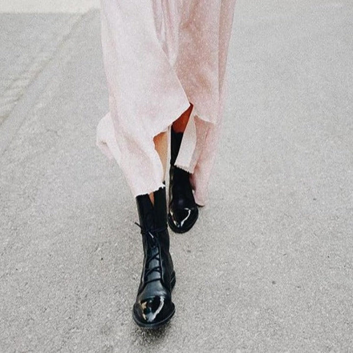 Bottines cheville pour femme en cuir noir à finition lisse et grainée, avec laçage sur le devant, bout arrondi et semelle plate robuste, portées en extérieur avec une robe fluide claire.