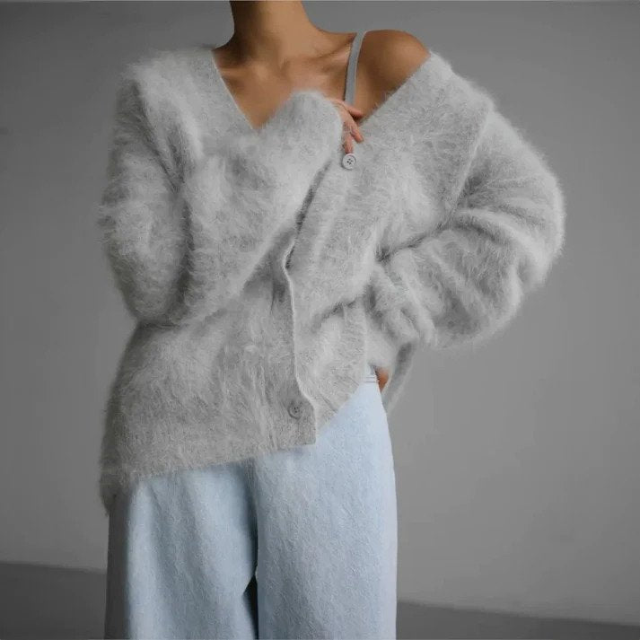Femme portant un cardigan duveteux gris clair légèrement ouvert, laissant apparaître une bretelle, associé à un pantalon ample de couleur pastel, dans un décor minimaliste et moderne.
