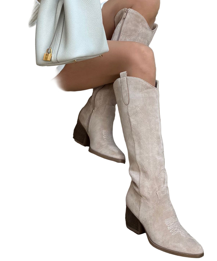 Jambes d’une femme assise sur des marches, portant des bottes hautes en daim beige à talons blocs, au style western moderne, assorties à une tenue claire et un sac élégant en cuir. Les bottes présentent des détails brodés subtils et une silhouette tendance, idéale pour un look chic et contemporain.