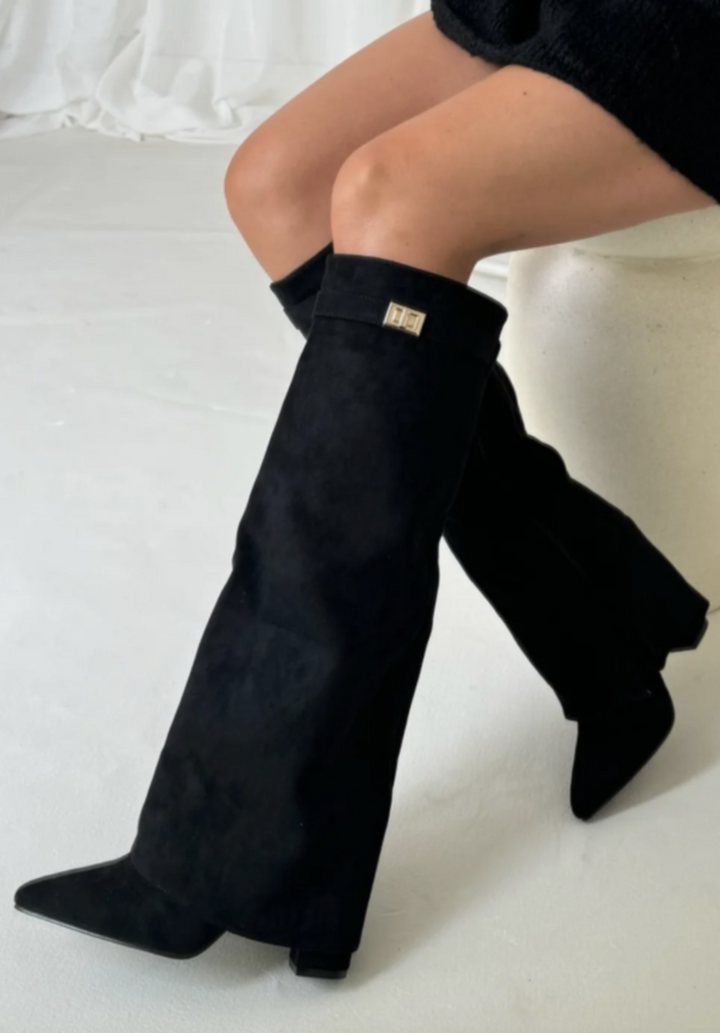Bottes noires à bout pointu avec revers et détail métallique, talon moyen, portées en position assise, vue de côté.