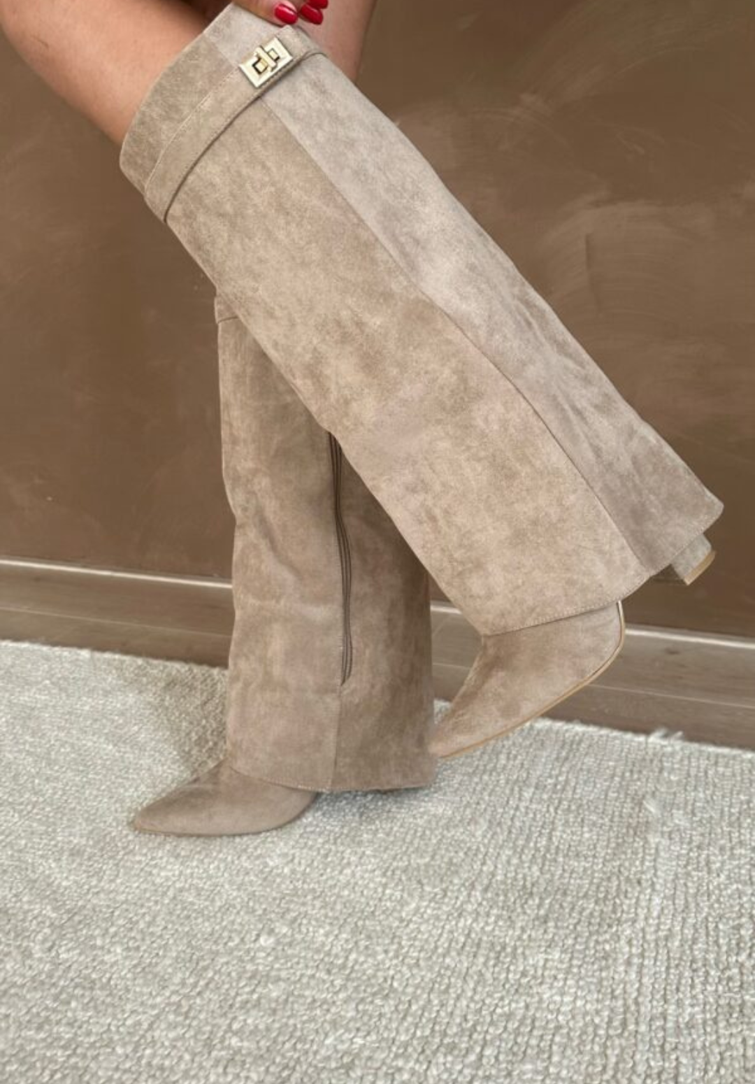 Bottes taupe effet daim à bout pointu avec fermeture éclair latérale et boucle dorée, talon moyen, portées jambes croisées sur tapis.