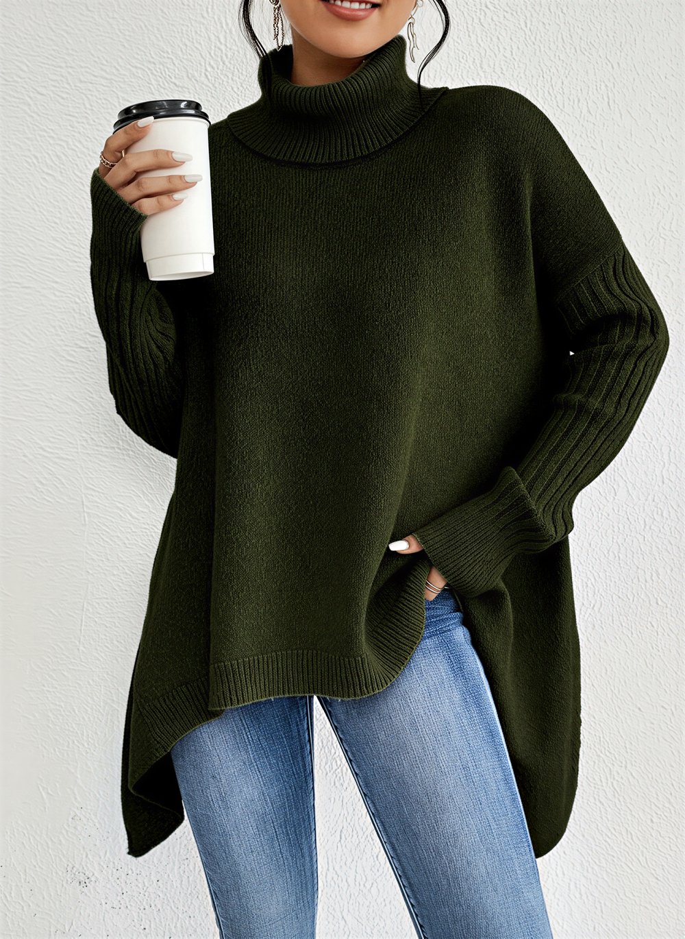 Une femme porte un pull à col roulé vert ample, à manches longues côtelées et à ourlet asymétrique. Elle tient une tasse de café à la main et porte un jean bleu slim, créant ainsi un look décontracté et élégant, parfait pour les mois les plus froids.