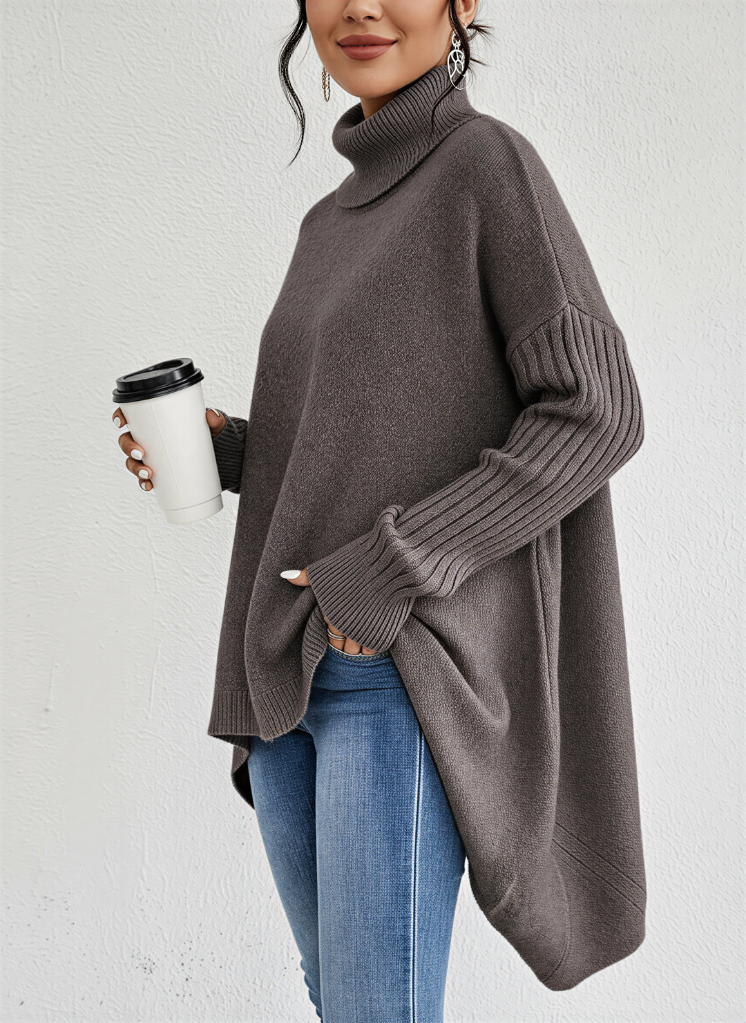 Femme portant un pull à col roulé ample de couleur taupe, avec des manches longues côtelées et une coupe asymétrique. Elle tient une tasse à café et porte un jean bleu, créant une allure chic et décontractée, parfaite pour l’automne et l’hiver.