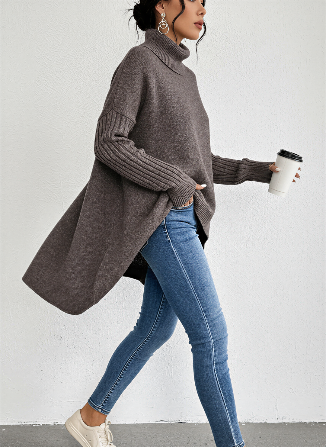 Femme marchant avec un café à la main, vêtue d’un pull à col roulé marron à coupe ample et fluide. Le vêtement présente des manches longues côtelées et un ourlet asymétrique allongé à l’arrière, associé à un jean bleu ajusté et des baskets blanches pour un look moderne, confortable et décontracté.