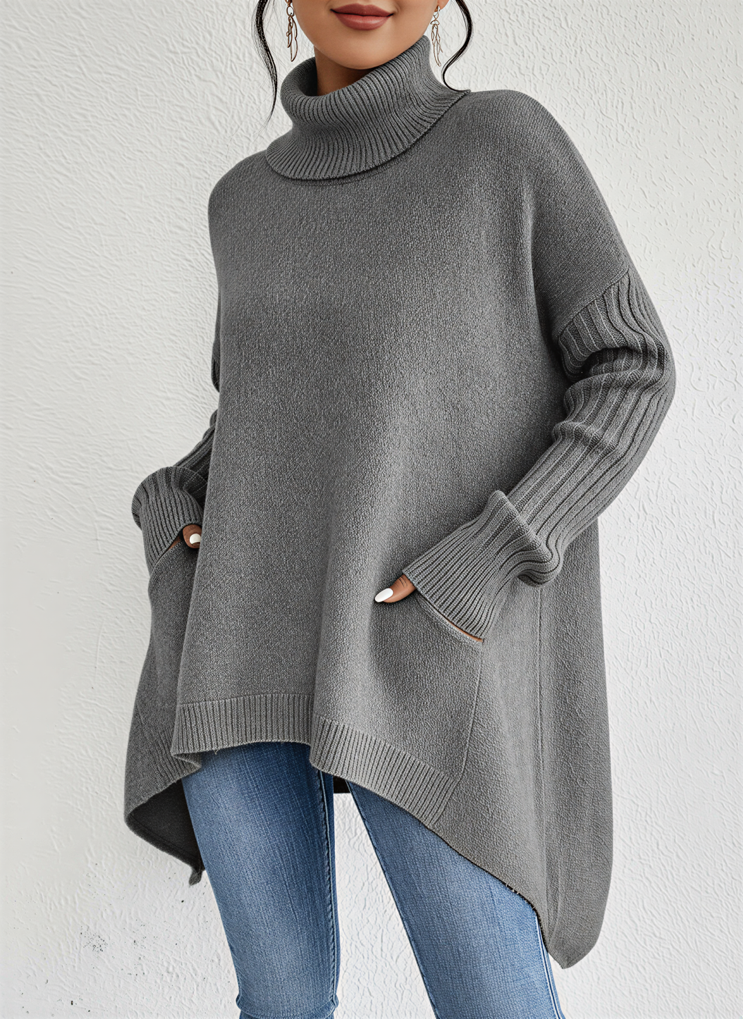Femme portant un pull à col roulé gris, coupe ample et moderne, avec des manches longues côtelées et deux poches latérales. Le vêtement présente une base asymétrique et est associé à un jean bleu pour un style chic, minimaliste et confortable idéal pour les journées fraîches.