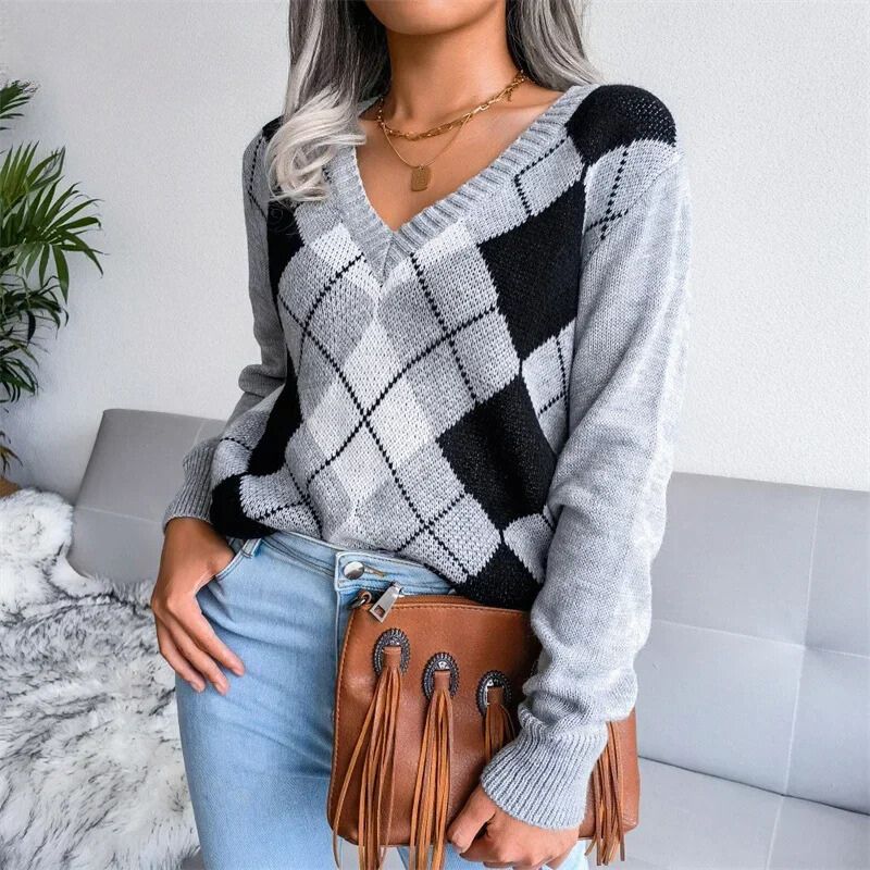 Une femme porte un pull à col en V en maille douce, orné d’un motif à losanges de style argyle dans les tons gris, noir et blanc. Le pull présente une coupe légèrement ample avec des manches longues et des bords côtelés. Elle l’associe à un jean bleu clair et un sac à main marron à franges, créant une tenue hivernale élégante et moderne.