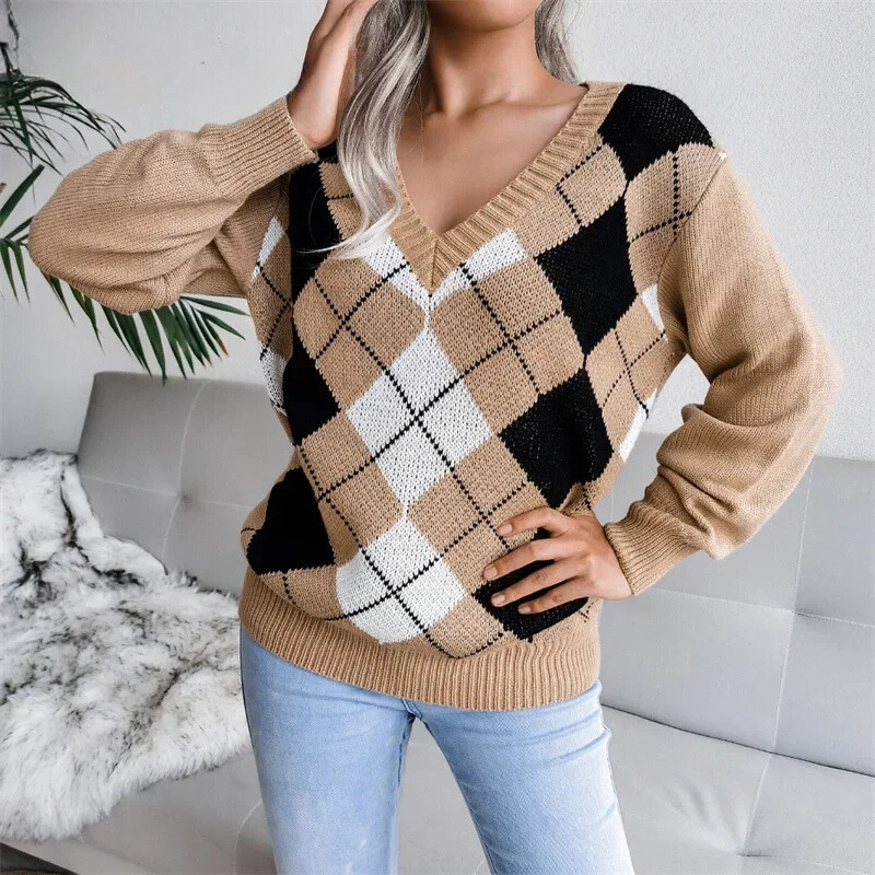 Une femme porte un pull en maille à col en V avec un motif à losanges de style argyle dans les tons beige, noir et blanc. Le pull présente une coupe décontractée avec des manches légèrement bouffantes et des bords côtelés. Il est associé à un jean bleu clair, créant une tenue confortable et élégante idéale pour la saison hivernale.