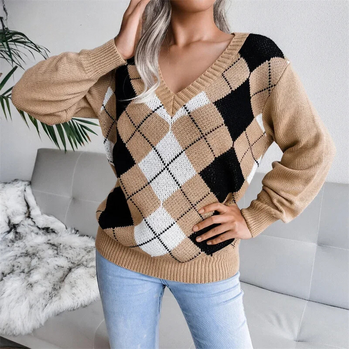 Une femme porte un pull en maille à col en V avec un motif à losanges de style argyle dans les tons beige, noir et blanc. Le pull présente une coupe décontractée avec des manches légèrement bouffantes et des bords côtelés. Il est associé à un jean bleu clair, créant une tenue confortable et élégante idéale pour la saison hivernale.