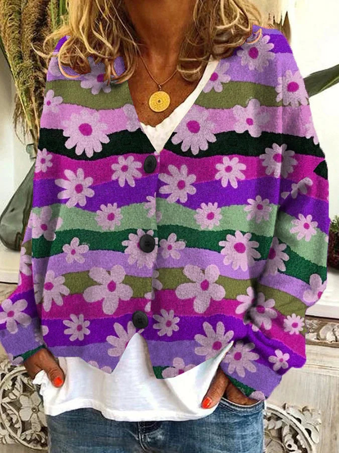 Femme portant un cardigan ample à imprimé fleuri dans les tons violets, verts et roses, avec de grands boutons noirs sur le devant. Le cardigan présente un motif de marguerites blanches sur des bandes ondulées colorées, créant un style rétro et original. La tenue est complétée par un t-shirt blanc et un jean décontracté, pour un look vintage et confortable.
