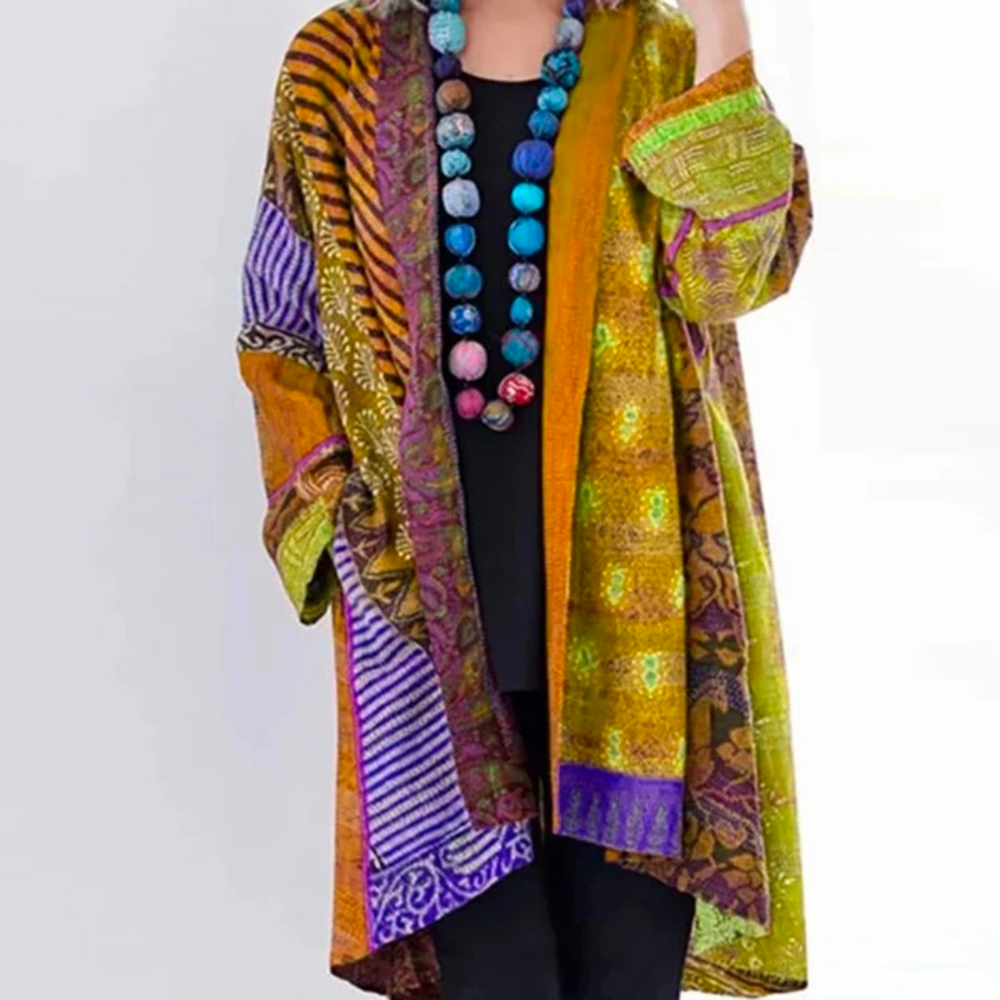 Femme portant un cardigan long et fluide au style tendance, avec motifs norvégiens multicolores dans des tons chauds de jaune, orange, vert et violet, manches longues et coupe ample, porté ouvert sur une tenue sombre pour une allure bohème et artistique.